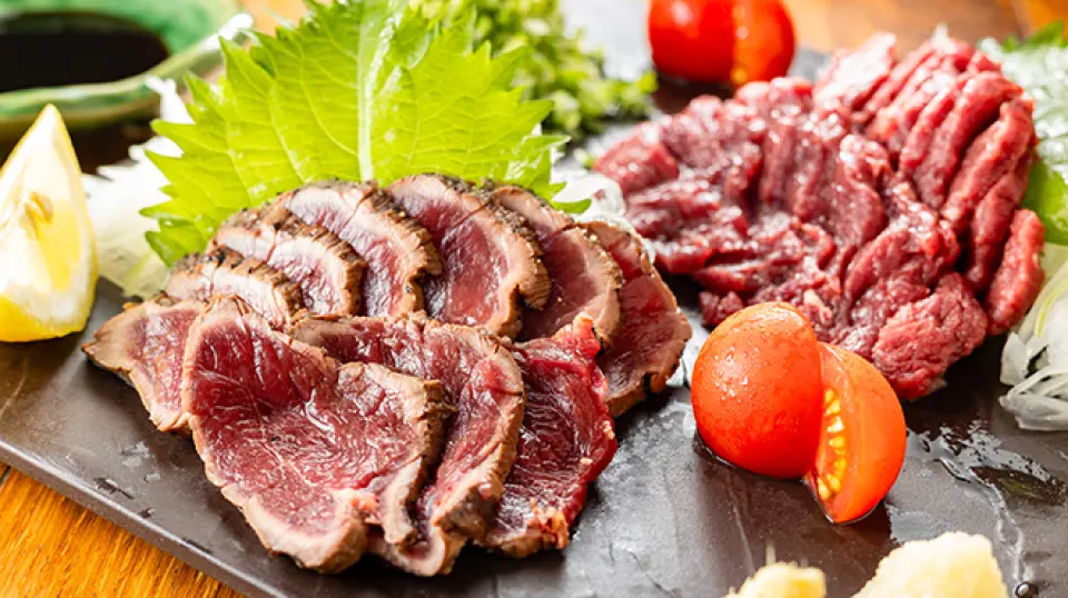 Thinly sliced seared meat tataki garnished with shiso leaves, fresh lemon, and cherry tomatoes served on a dark slate plate in Tokyo.