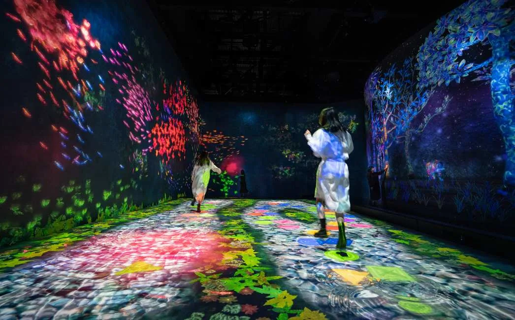 People walking through an immersive digital art installation featuring glowing floor patterns and walls projected with vibrant schools of fish and colorful forest scenes