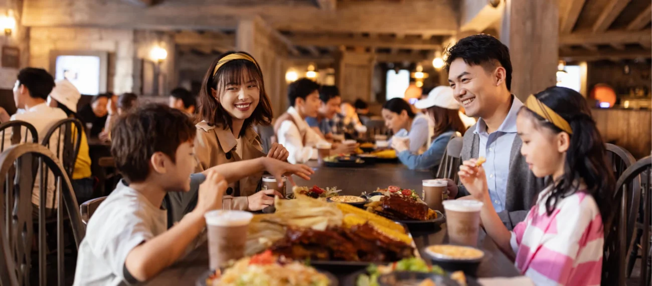 A family enjoying a meal together in the rustic, wood-beamed dining hall of the Three Broomsticks restaurant in Hogsmeade.