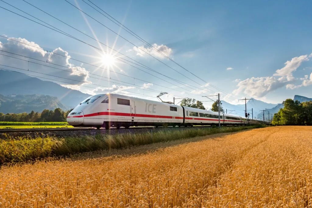 A white German ICE high-speed train travels through a golden wheat field under a bright sun and blue sky.