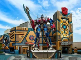 A towering statue of Optimus Prime standing in front of the Transformers Metrobase attraction building under a bright blue sky.