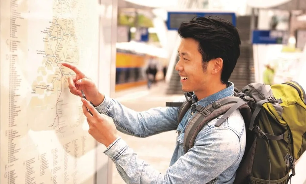A smiling man with a green backpack points at a public transit map while holding a smartphone at a station.