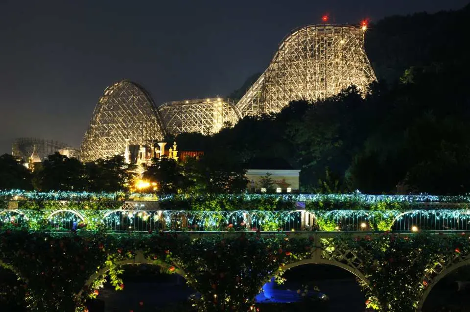 The illuminated wooden structure of the T Express roller coaster shining against the night sky above a bridge adorned with fairy lights at Everland.